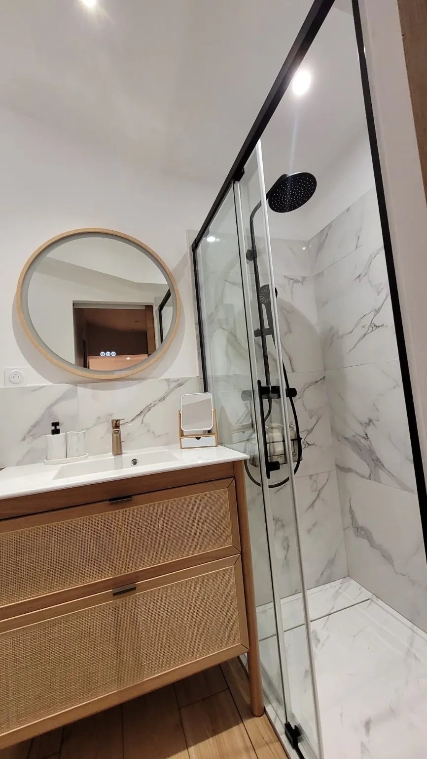 Meuble vasque en bois et cannage, miroir rond et douche avec paroi noire.